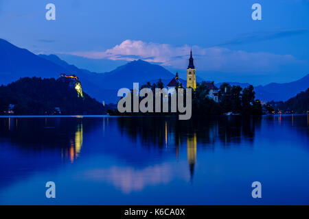 Illuminato isola di Bled, Blejski Otok, con la chiesa di pellegrinaggio dedicata all Assunzione di Maria si vede attraverso il lago di Bled e blejsko jezero di notte Foto Stock