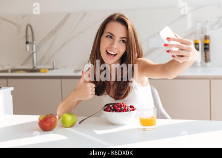 Sorridente bella donna prendendo un selfie e mostrando i pollici fino gesto pur avendo una sana prima colazione nella cucina Foto Stock