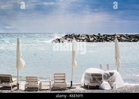 Tre piegato ombrelloni e sdraio sulla spiaggia con mare tempestoso in background Foto Stock