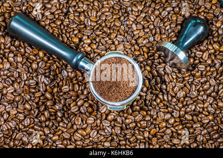 Espresso il concetto: il caffè macinato in una macchina per caffè espresso portafilter cestello, con antimanomissione in acciaio a fianco, sulla parte superiore di un sacco di chicchi di caffè. Foto Stock