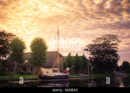 Una barca a vela olandese tradizionale barca ancorata in un canale esterno di una casa con la golden cielo nuvoloso, Holland, Paesi Bassi. Foto Stock