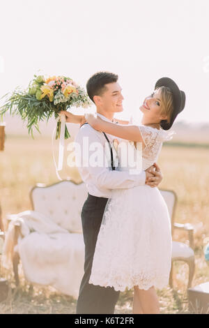 La felice sposi novelli sono abbracciando in background del divano posto nel soleggiato campo. La sposa è in possesso della enorme colorati mazzo Foto Stock