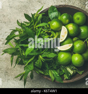 Flatlay di appena raccolti organici di lime e foglie di menta per la realizzazione di cocktail o limonata in piatto di legno su cemento grigio sfondo di pietra, vista dall'alto, Foto Stock