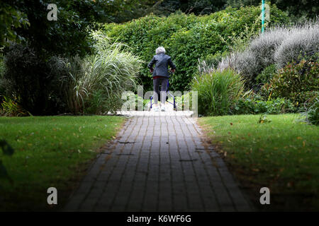 Una donna anziana foto camminando lentamente con un tri walker aiuto attraverso un parco in felpham, west sussex, Regno Unito. Foto Stock