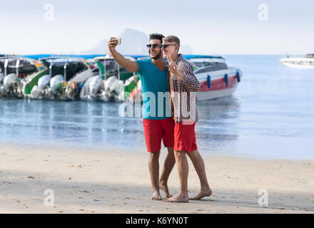 Turisti uomo giovane tenendo selfie foto nella parte anteriore della barca dalla coda lunga sulla spiaggia sul cellulare smart phone, due giovani ragazzi sorridenti sul mare vacanza Foto Stock