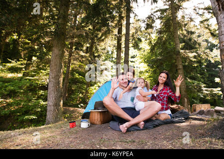 Giovane e bella famiglia con graziosi piccole figlie camping in foresta, seduti nella parte anteriore della tenda. Foto Stock