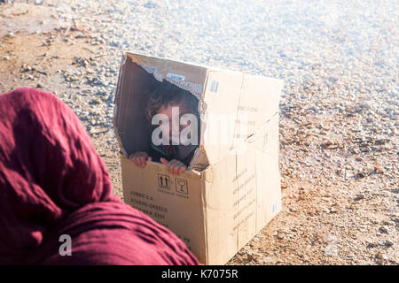 Un bambino gioca felicemente in una scatola di cartone sul fosco pietraie di ritsona Refugee Camp in Grecia. madre {primo piano) è nascosto nel velo. Foto Stock