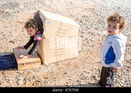 Un bambino gioca felicemente in una scatola di cartone sul fosco pietraie di ritsona Refugee Camp in Grecia come il suo amico guarda la telecamera. Foto Stock