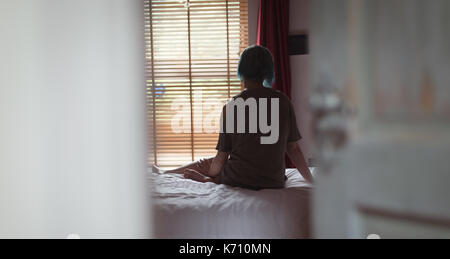 Vista posteriore della donna seduta dalla finestra sul letto di casa Foto Stock