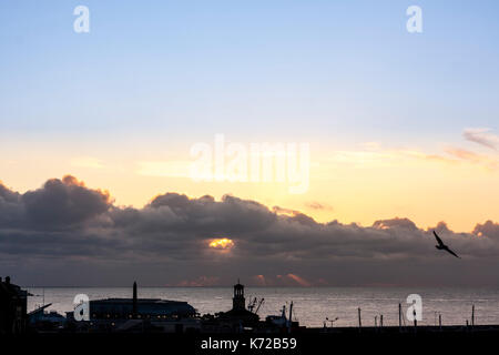 Inghilterra, Ramsgate. Sunrise visto attraverso la nube spessa appeso nello strato sull'orizzonte sul mare, sul Canale Inglese. Gli edifici in primo piano in silhouette. Seagull battenti. Foto Stock