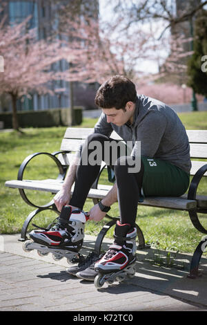 Giovane uomo che indossa i pattini a rotelle durante la seduta sul banco a park durante la giornata di sole Foto Stock