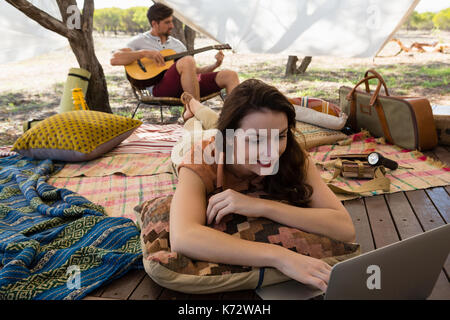 Donna rilassato utilizzando notebook con il giovane a suonare la chitarra in tenda Foto Stock