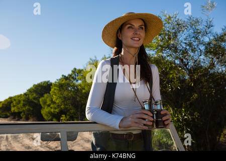 Sorridente giovane donna tenendo il binocolo che guarda lontano mentre in piedi in fuoristrada Foto Stock