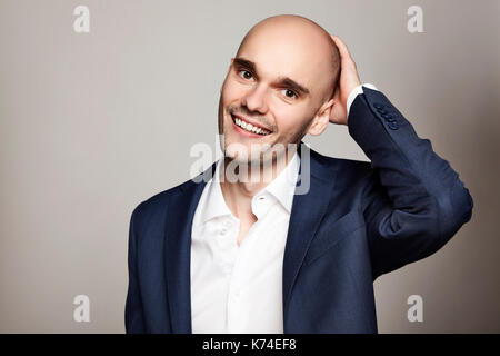 Close-up ritratto di una giovane uomo calvo accarezzare la sua testa. sta sorridendo. sfondo grigio. orizzontale. Foto Stock