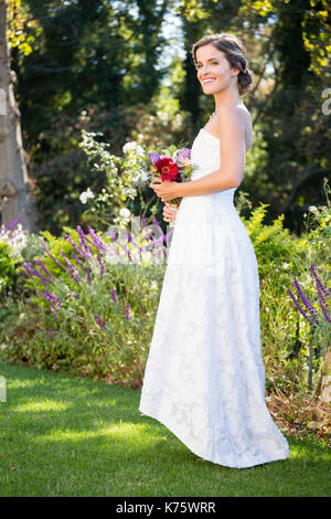 Sorridente sposa bouquet di contenimento mentre si sta in piedi sul campo erboso in cantiere Foto Stock