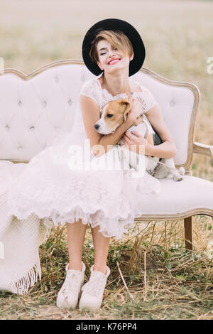 Il Sorridente vintage-sposa vestito con il cappello nero è abbracciando il bel cane e seduto sul divano nel soleggiato Campo. Foto Stock