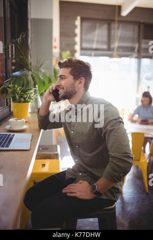 Vista laterale di sorridere giovane uomo parlando al cellulare mentre è seduto al banco in coffee shop Foto Stock