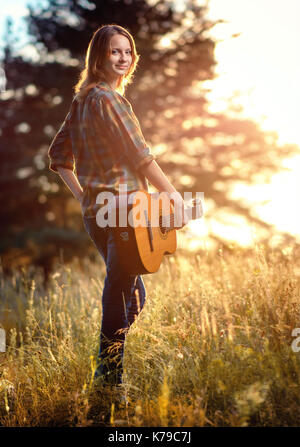 Ragazza sorridente in un plaid shirt con una chitarra in mano guarda oltre la sua spalla per la telecamera. Una passeggiata nel parco di autunno al tramonto. Foto Stock