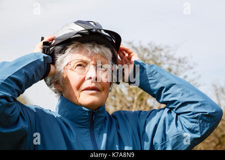 Active senior donna anziana pensionato mettendo su una bicicletta casco preparando per andare a fare un giro in bicicletta per mantenersi in forma di pensionamento. Inghilterra, Regno Unito, Gran Bretagna Foto Stock