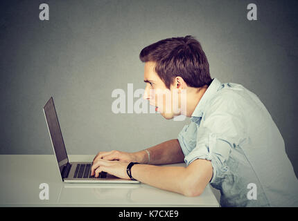 Scioccato perplesso uomo seduto nella parte anteriore del computer portatile che stanno guardando sullo schermo isolato sul muro grigio Sfondo. faccia emozione espressione di sentimenti Foto Stock