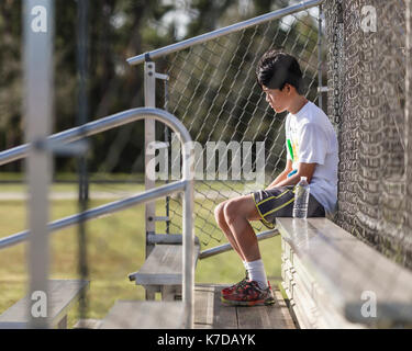 Riflessivo ragazzo adolescente seduto sulla panchina contro opaco recinzione al campo da gioco Foto Stock