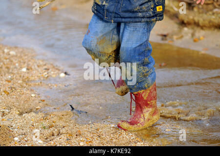 Un bambino o un ragazzo che indossa red wellies stivali da pioggia e coperto di fango e bagnato con acqua attraverso il salto in pozzanghere sotto la pioggia e il tempo umido in inverno Foto Stock