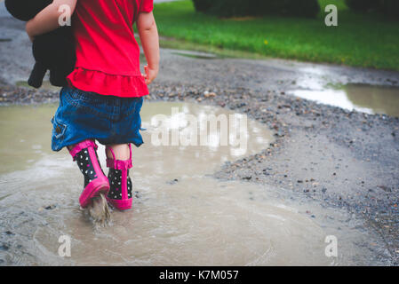 Un bambino a camminare in una pozza di fango in stivali da pioggia portando un peluche Foto Stock