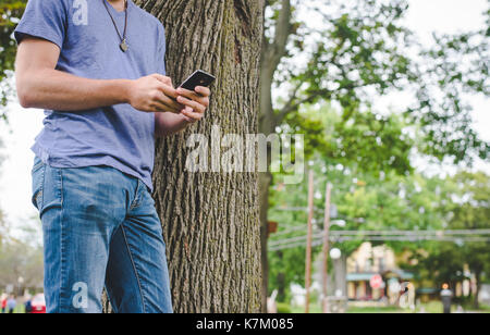 Un giovane uomo guarda al suo cellulare mentre appoggiata contro un albero. Foto Stock