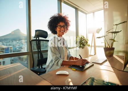 Donna sorridente designer seduto alla sua scrivania. Afro American giovane donna al suo posto di lavoro creativo. Foto Stock
