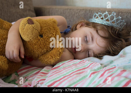 Close-up verticale bambina dormire nel letto. ragazza con una corona di principessa sul suo capo nel letto che abbraccia un orsacchiotto giocattolo, concetto di sogni tranquilli Foto Stock