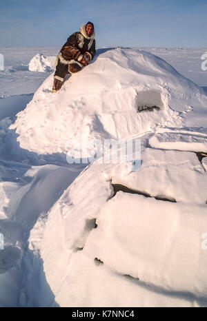 Inuit uomo anziano, metà 60s, vestito di Caribou Coffee Company tradizionale abbigliamento di pelle, pone sulla cima di un tradizionale igloo. igloo effettiva non sono lasciati con blocchi mostrante. Quando i blocchi sono a posto, l'igloo è coperto di neve per un ulteriore isolamento. Foto Stock