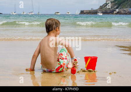 Un ragazzo con giocattoli da spiaggia siede su un litorale osservando le onde Foto Stock