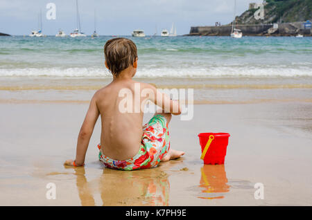 Un ragazzo con giocattoli da spiaggia siede su un litorale osservando le onde Foto Stock