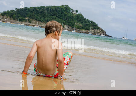 Un ragazzo in un costume da bagno si siede su un litorale osservando le onde Foto Stock