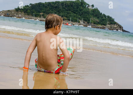 Un ragazzo in un costume da bagno si siede su un litorale osservando le onde Foto Stock