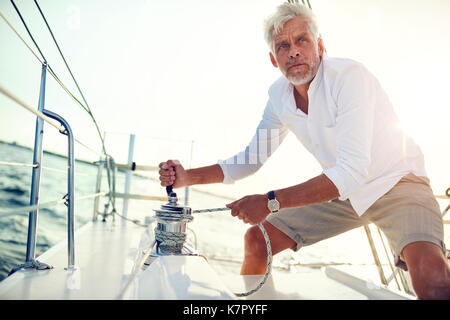 Uomo maturo in piedi sul ponte della barca di un avvolgimento di un argano mentre sono fuori per una vela di un pomeriggio soleggiato Foto Stock