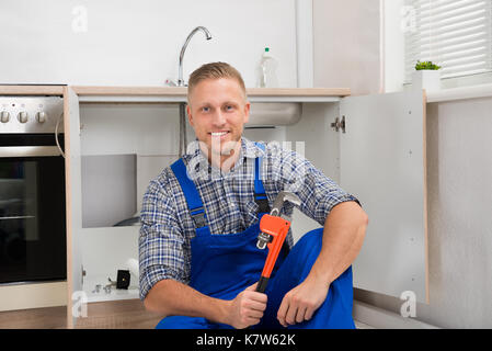 Felice giovane idraulico con una chiave regolabile seduto in camera con cucina Foto Stock