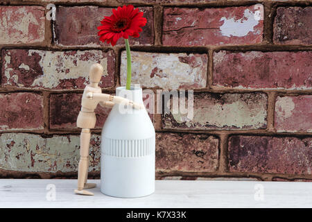 Manichino di legno con un vaso e fiore rosso contro un muro di mattoni in background. Foto Stock