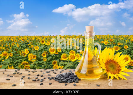 Olio di semi di girasole in una bottiglia di vetro, pila di semi di girasole, girasoli freschi sul tavolo di legno con sfondo naturale. blooming campo di girasoli con blue s Foto Stock