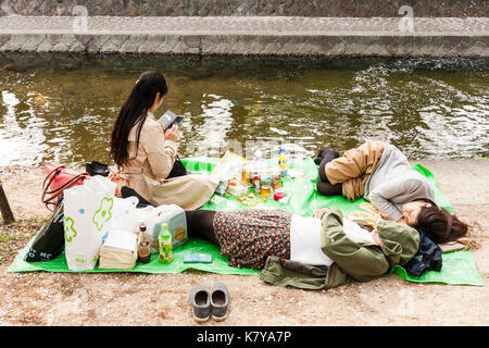 Molla tradizionale fiore di ciliegio parte dal fiume Shukugawa, nishinomiya, Giappone. Due giovani donne addormentato sul tappeto verde, una terza seduta di lettura. Foto Stock