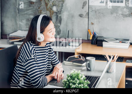 Giovani asiatici imprenditrice casual braccio sulla scrivania resto pongono con computer portatile e ascolto di musica attraverso le cuffie e guardando attraverso la finestra di ufficio ,lavoro Foto Stock