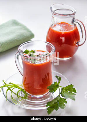 Il pomodoro succhi di verdura per la dieta e la pappa è versata in una tazza di vetro su una lastra di vetro e in un vetro trasparente caraffa con erbe su un bianco ba Foto Stock