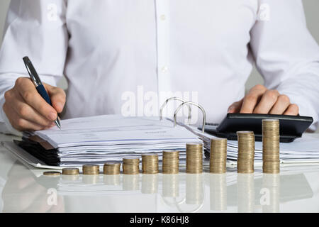 Sezione mediana di imprenditore calcolo fattura con monete impilate disposte a scrivania da ufficio Foto Stock