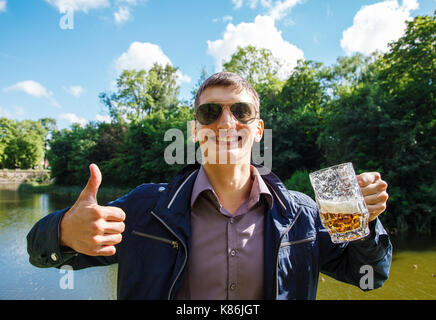Gioioso uomo sorridente tenendo un grande boccale di birra e che mostra i pollici fino all'aperto Foto Stock