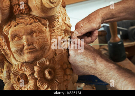 Close up di un artigiano artigiano, un intagliatore di legno lavorando su una polena di legno con un volto umano e forme floreali in un workshop, tenendo in mano un piccolo chris Foto Stock