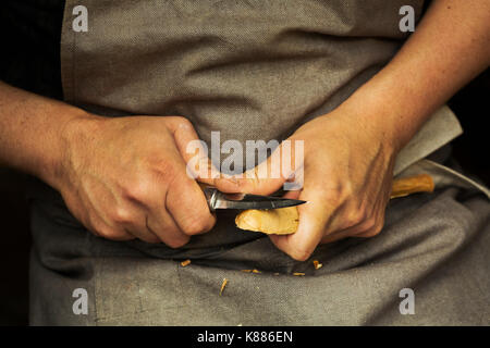 Close up di un artigiano di mani premendo e sagomatura di un piccolo pezzo di legno in un cucchiaio con una lama affilata lama, sagomando la ciotola all'indietro. Foto Stock