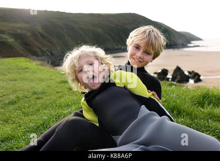 I fratelli giovani guardano alla telecamera come si gioca su erba pendio che si affaccia sulla spiaggia di indossare abiti bagnati. Foto Stock