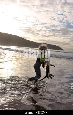 Giovane ragazzo sempre pronto a navigare con la sua tavola da surf in spiaggia indossando una muta su una giornata d'estate. Foto Stock