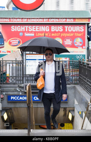 Maschio bianco indossando occhiali da sole emerge da una stazione della metropolitana di Londra con il suo ombrello aperto durante una doccia a pioggia. Foto Stock