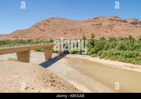 Bridge spanning oltre il letto asciutto del fiume con un po' d'acqua, le montagne e le palme in Marocco, Africa del nord. Foto Stock